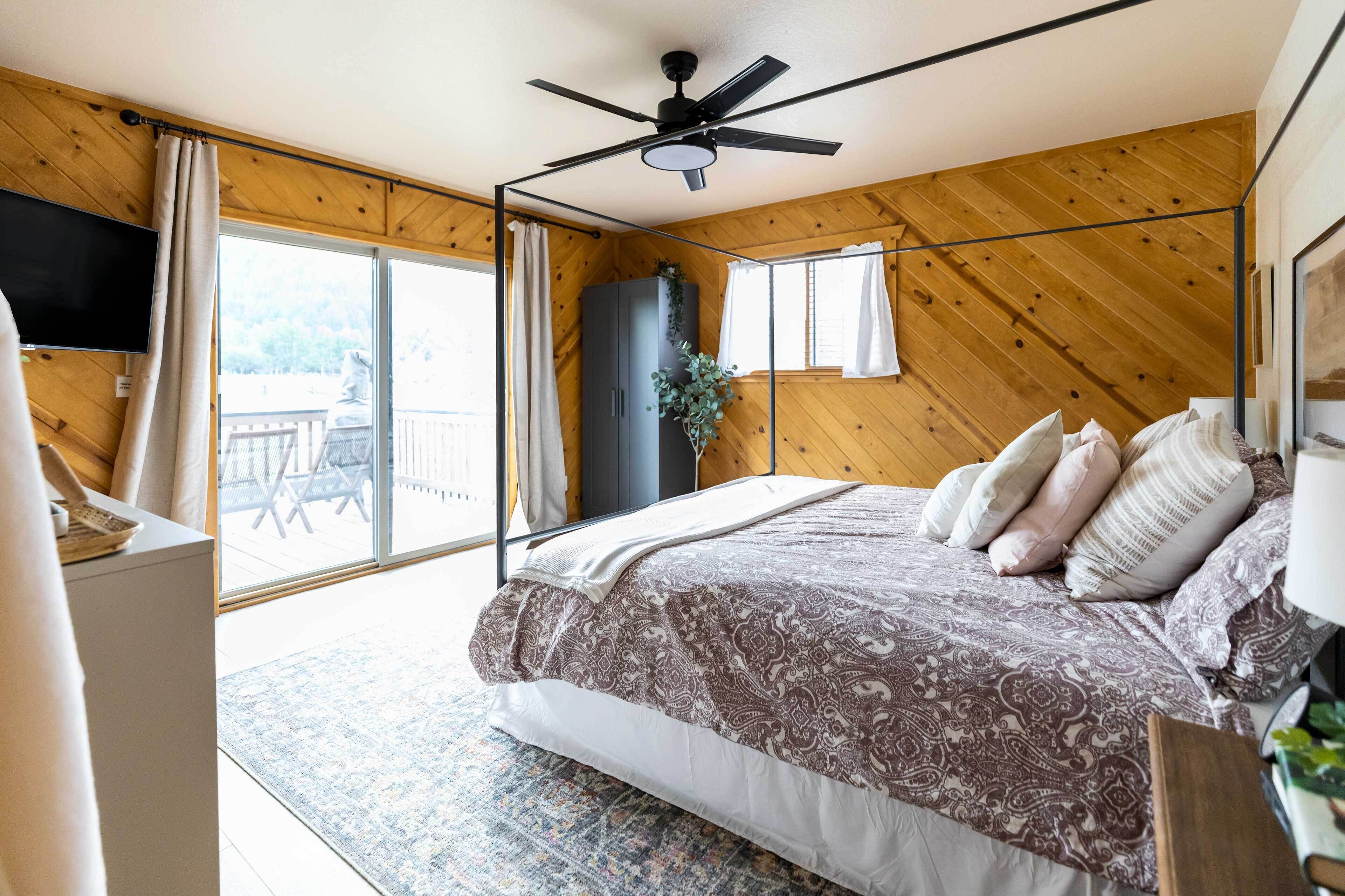 A bedroom with a bed, a ceiling fan, a large window, and a view of the outdoors.