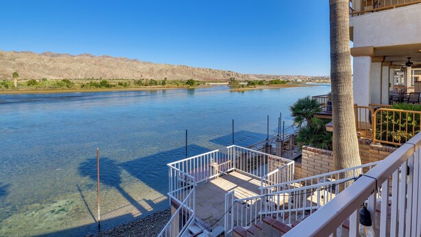 Vista de un río con cielo despejado, un balcón con mobiliario exterior y una palmera.