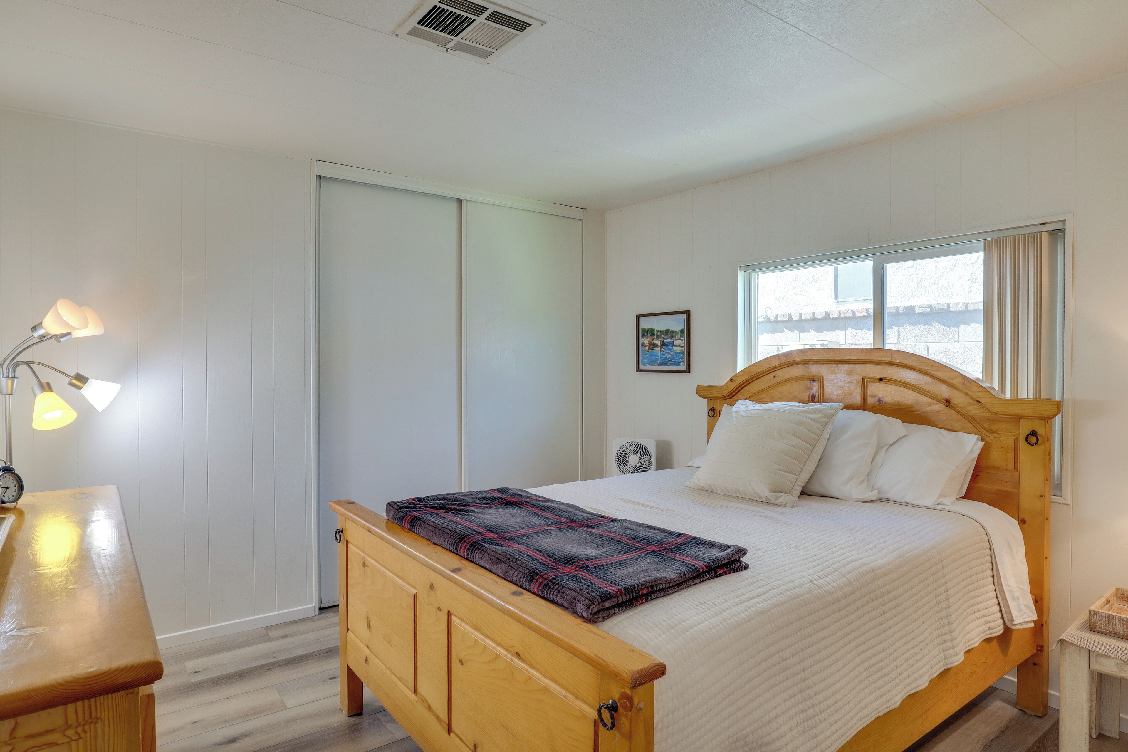 Un dormitorio con cama de madera, ropa de cama blanca, una mesita de noche con lámpara, una ventana con cortinas y un cuadro colgado en la pared.