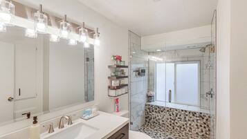 Un baño moderno con un espejo grande, un lavamanos blanco con dos grifos y una zona de ducha con mampara de cristal.
