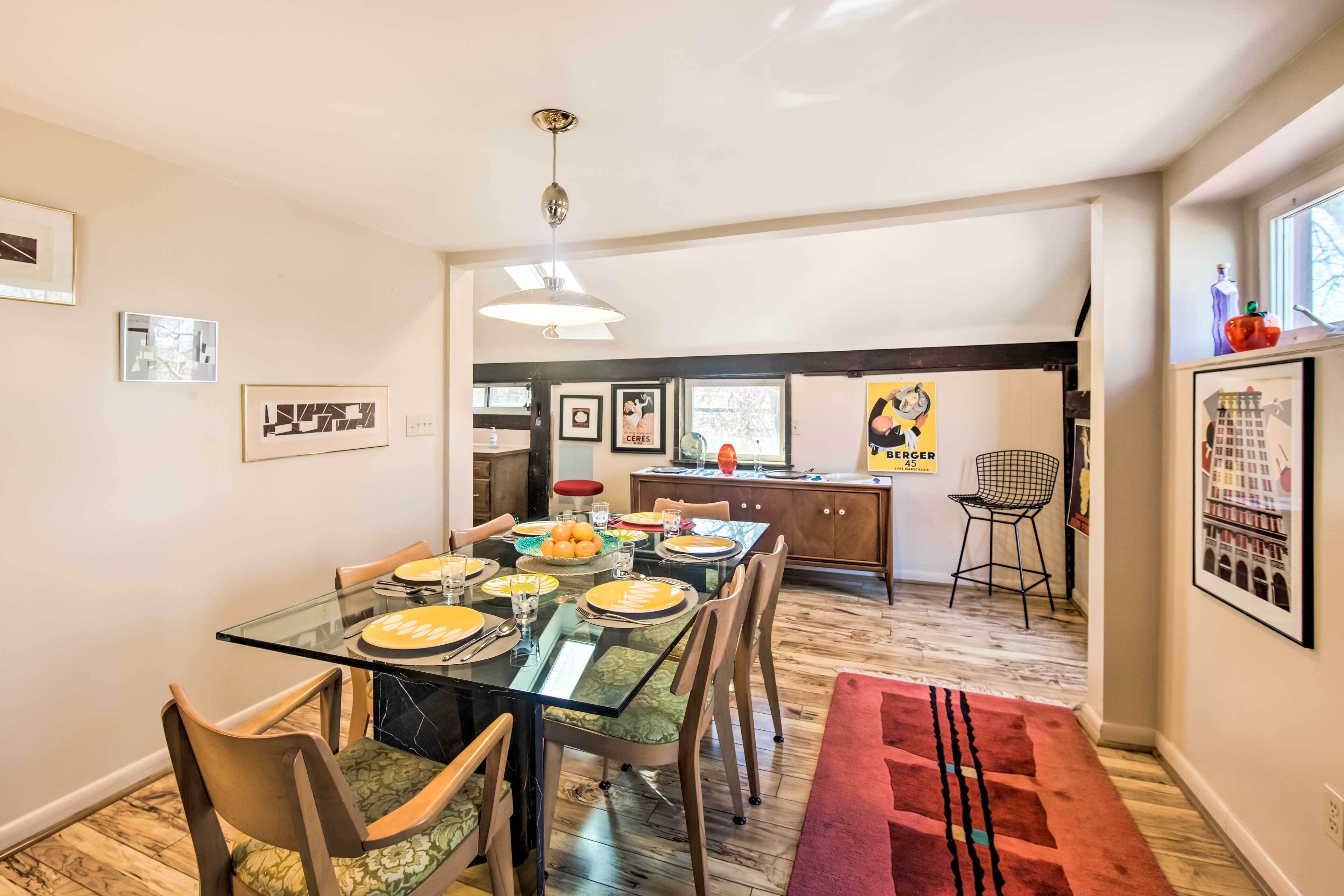A modern dining area with a glass table, chairs, and a red rug. There is a kitchen in the background with a sink and a chair.