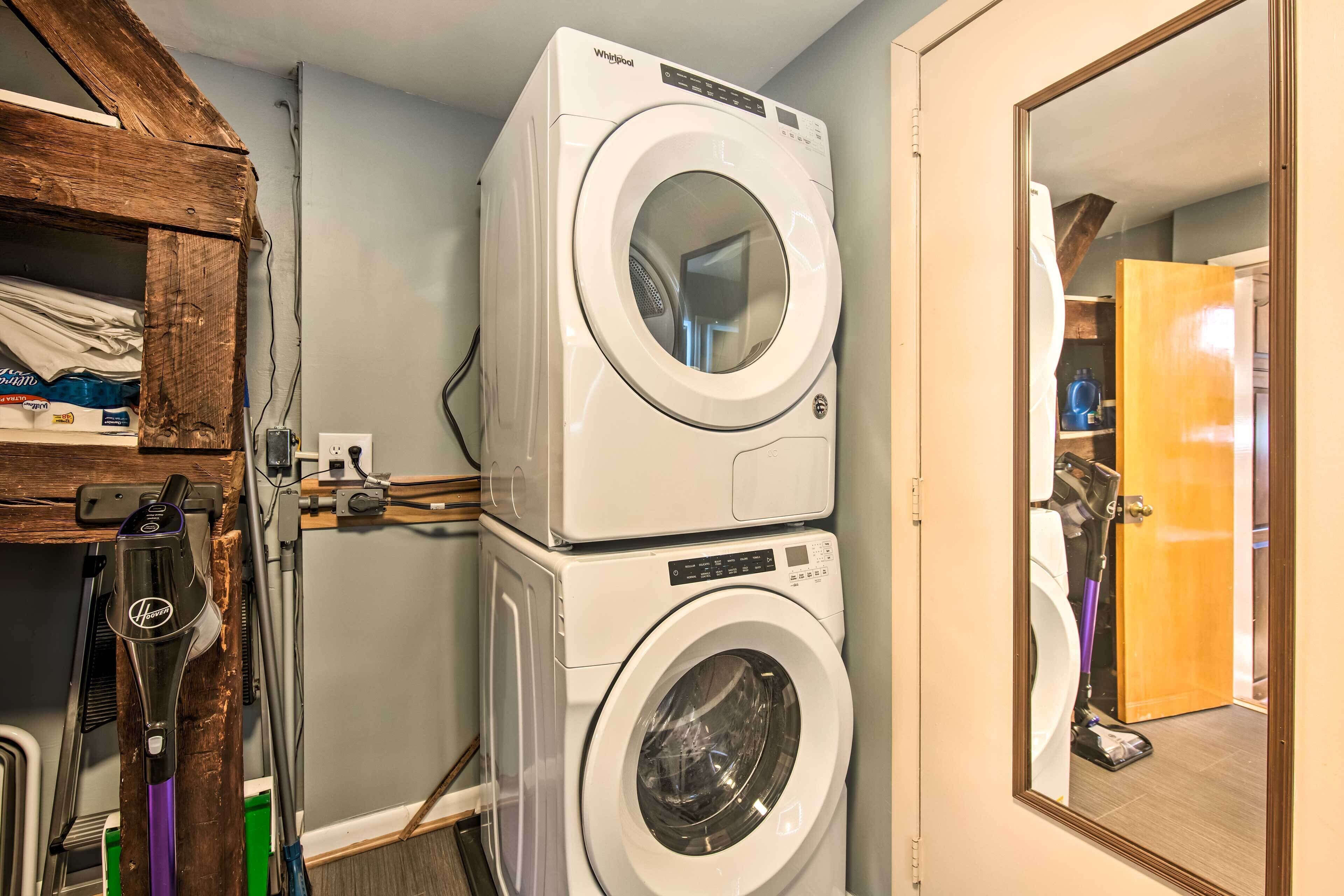 Un área de lavandería con lavadoras y secadoras apiladas, un estante de madera con productos de limpieza y un espejo que refleja la habitación.