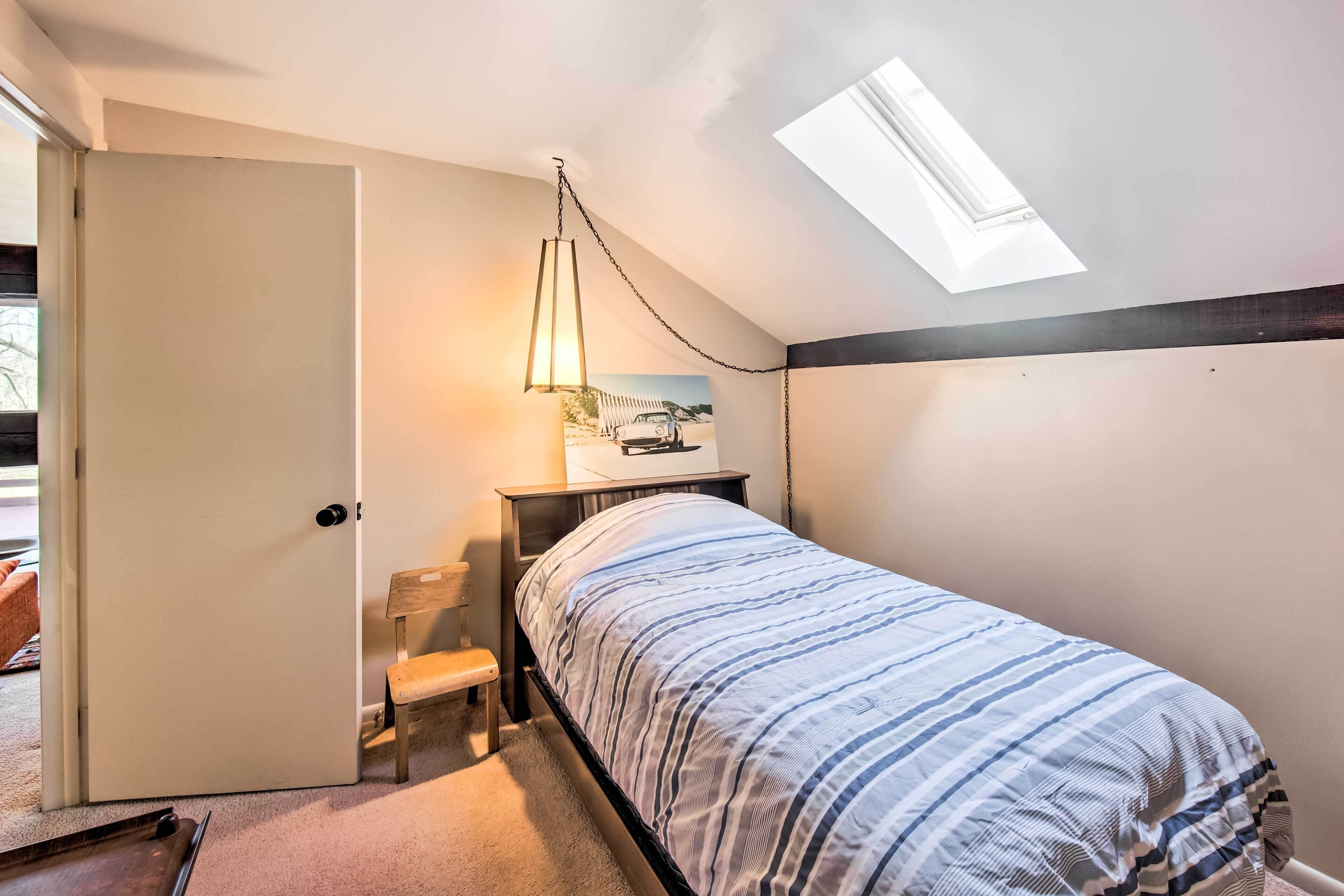 A bedroom with a bed, a nightstand, a chair, a small table, a painting, and a skylight.