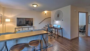 A modern living room with a wooden dining table, grey upholstered sofas, a large flat-screen TV, and a staircase leading to another level. - Cozy Cedar Rapids Pad - 2 Blocks to Mercy Care! (Cedar Rapids)