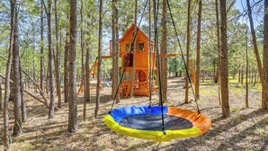 Une cabane en bois perchée dans un arbre, avec un trampoline, au milieu d’une forêt.