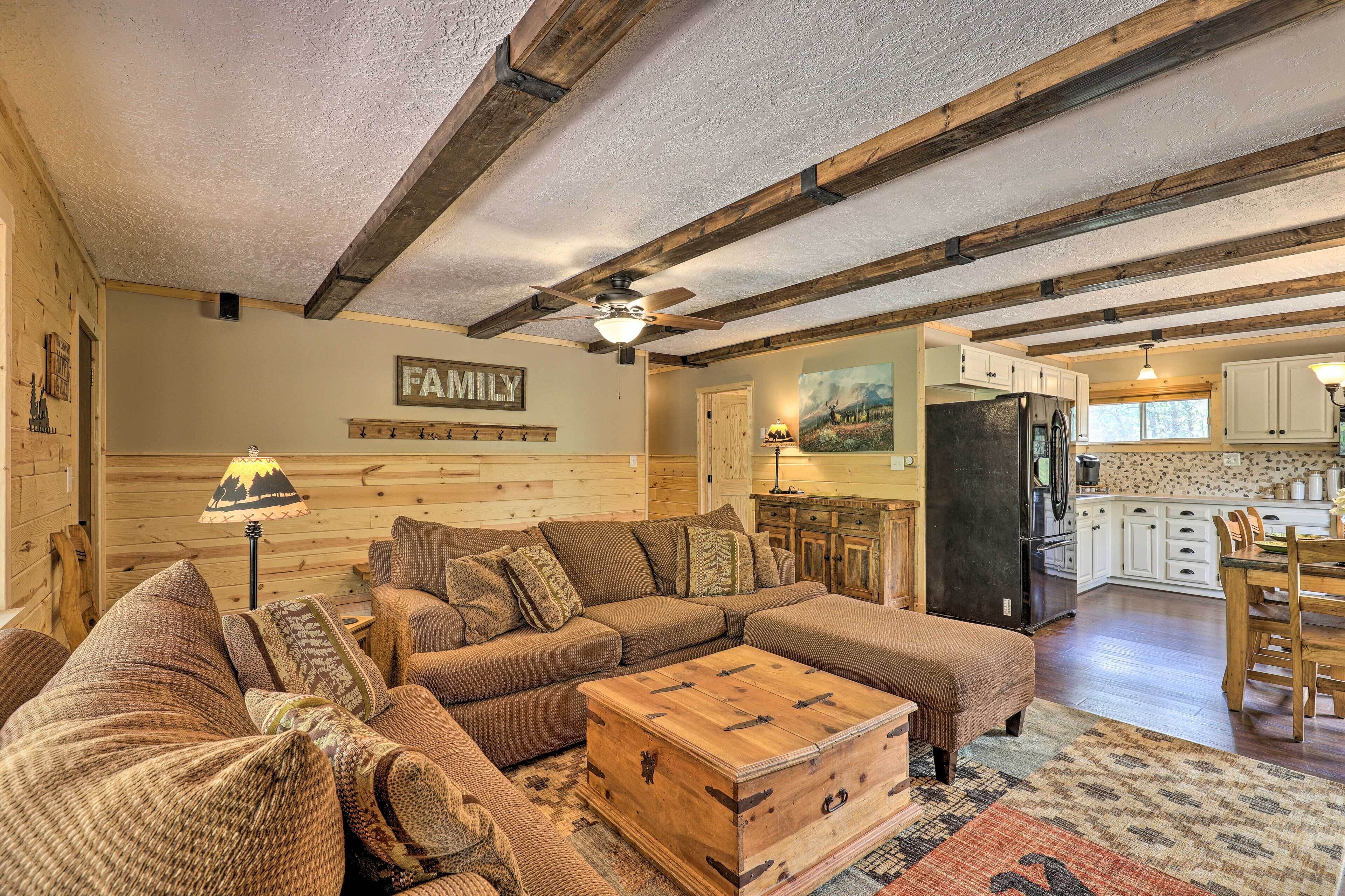 A cozy living room with a sectional sofa, a wooden coffee table, and a ceiling fan. A kitchen with white cabinets and a refrigerator is visible in the background.