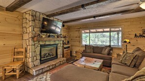 L’intérieur chaleureux d’un chalet avec une cheminée en pierre, des murs en bois, un téléviseur fixé au-dessus de la cheminée, un canapé et une grande fenêtre donnant sur le paysage extérieur.