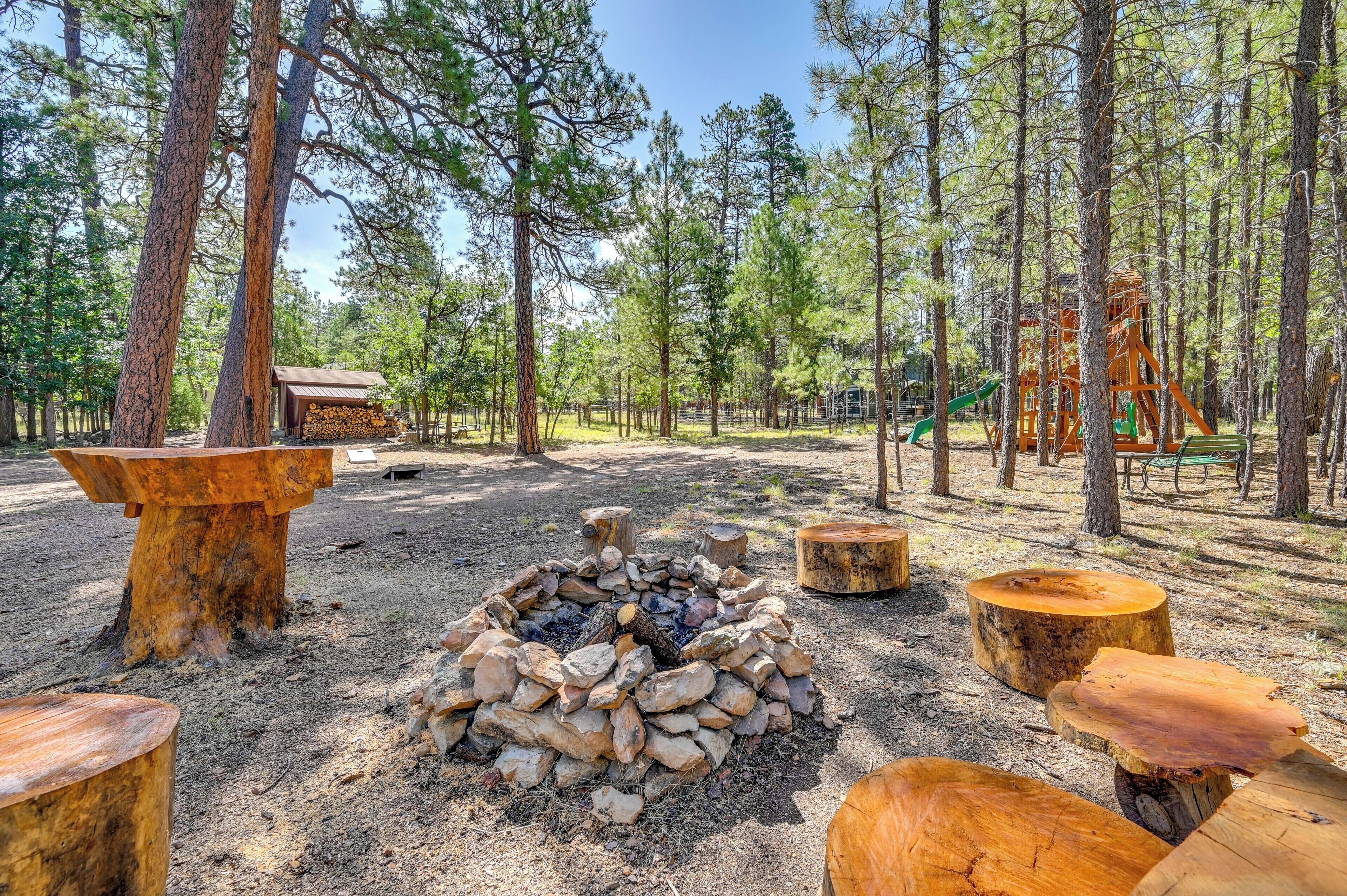 A forested campsite with a fire pit, wooden benches, and a playground structure.