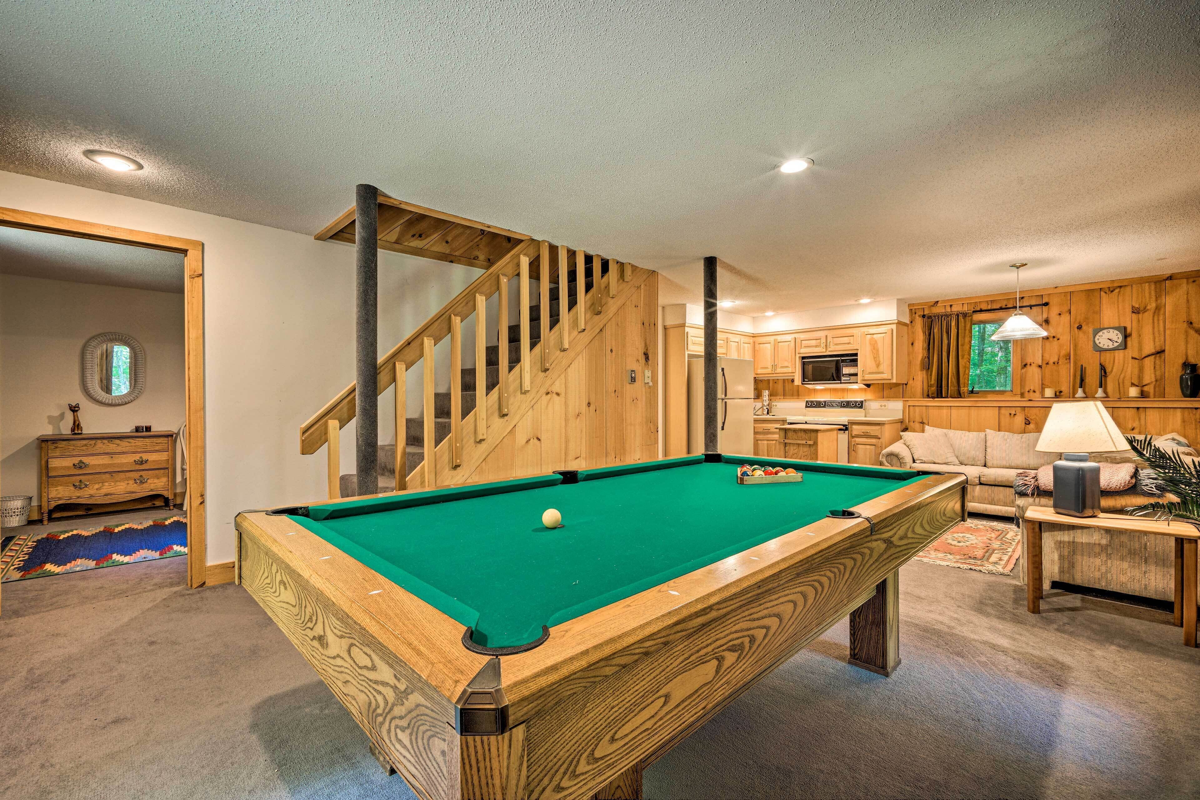 A pool table in a room with a wooden staircase, a kitchen area, and a living room with a sofa and lamp.