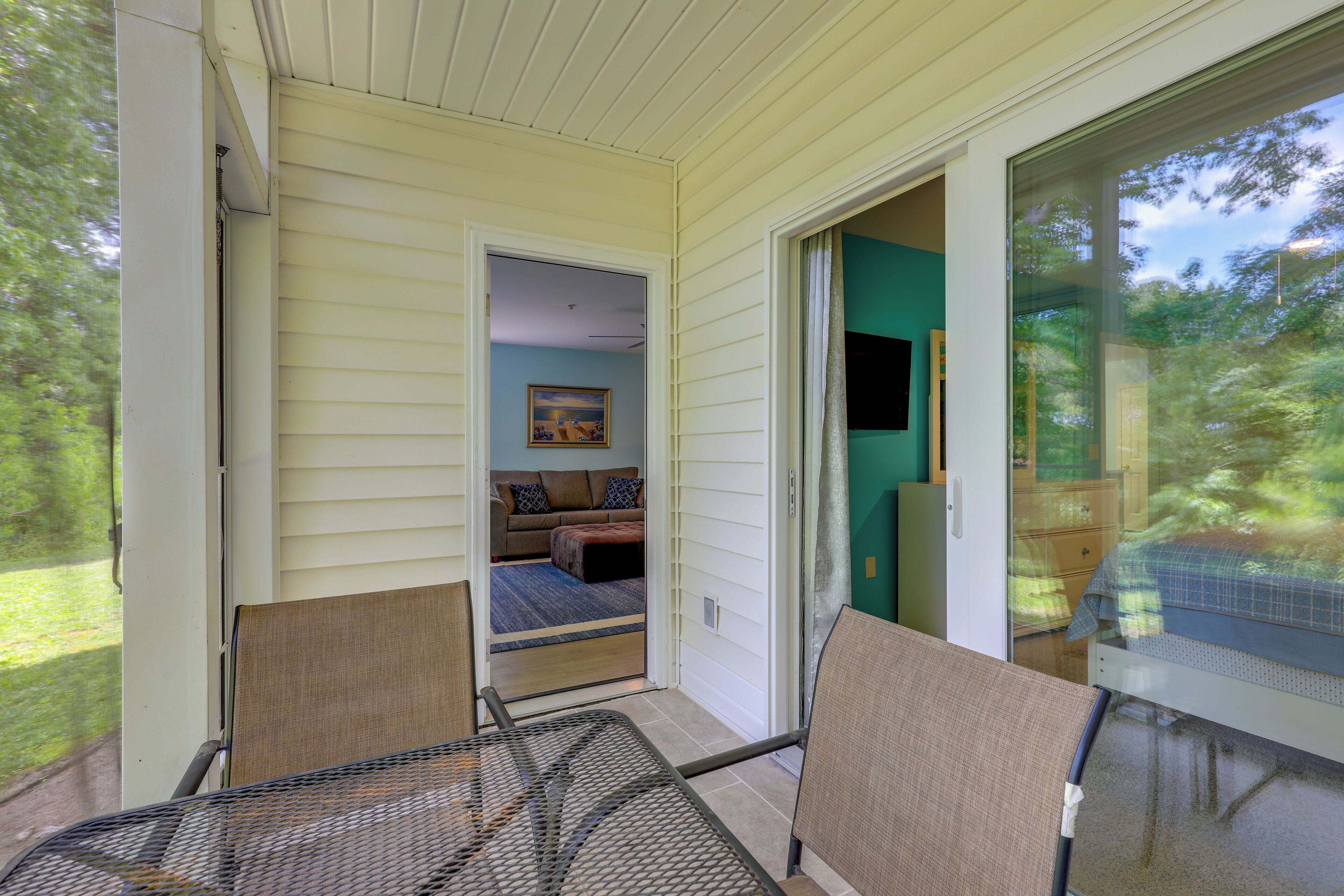 A covered porch with a sliding glass door leading to a bedroom with a bed and a painting on the wall.