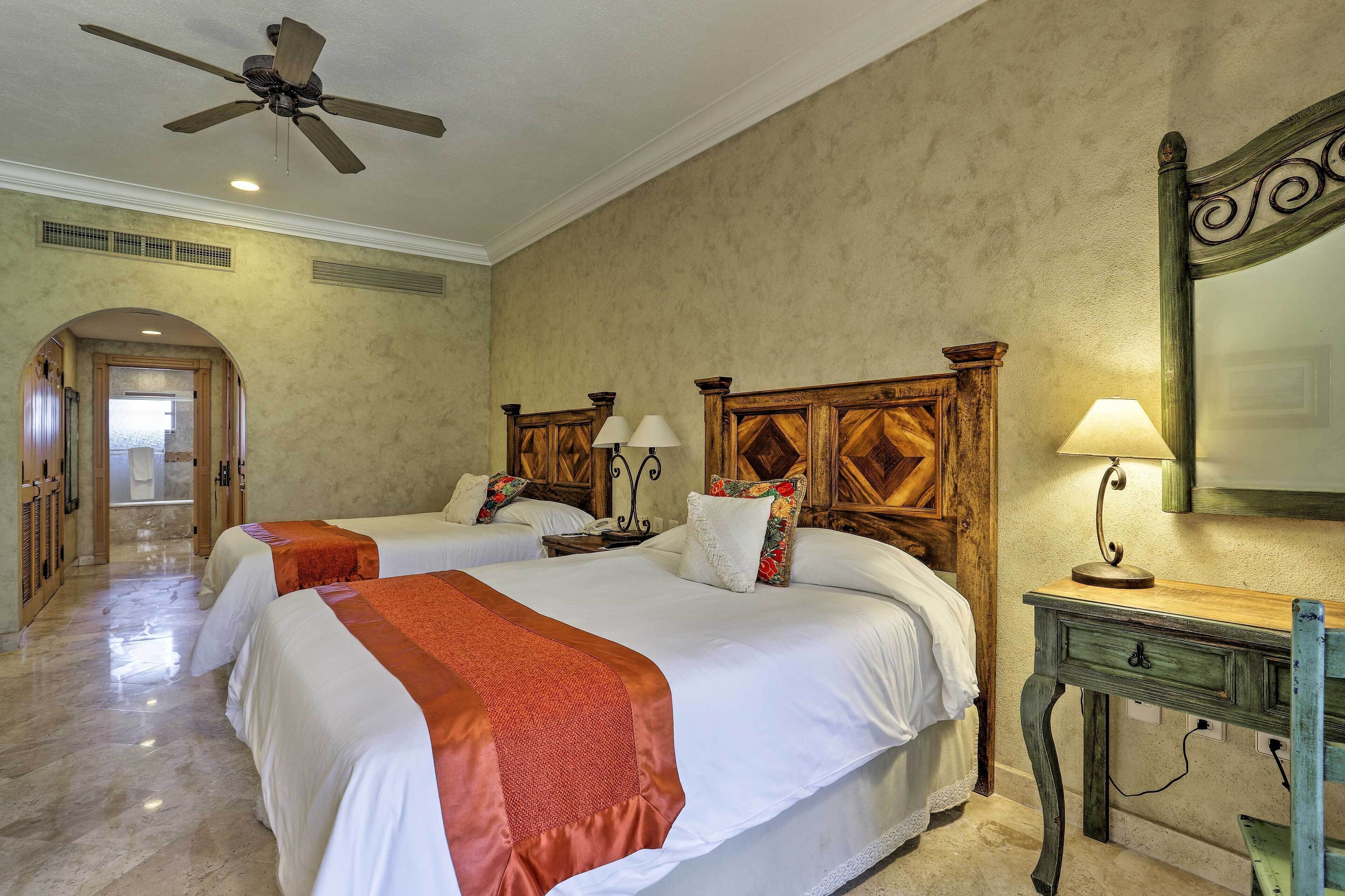 Ein Hotelzimmer mit zwei Betten, einem Deckenventilator, einem Holzkopfende, einem Nachttisch mit Lampe und einem Spiegel an der Wand.