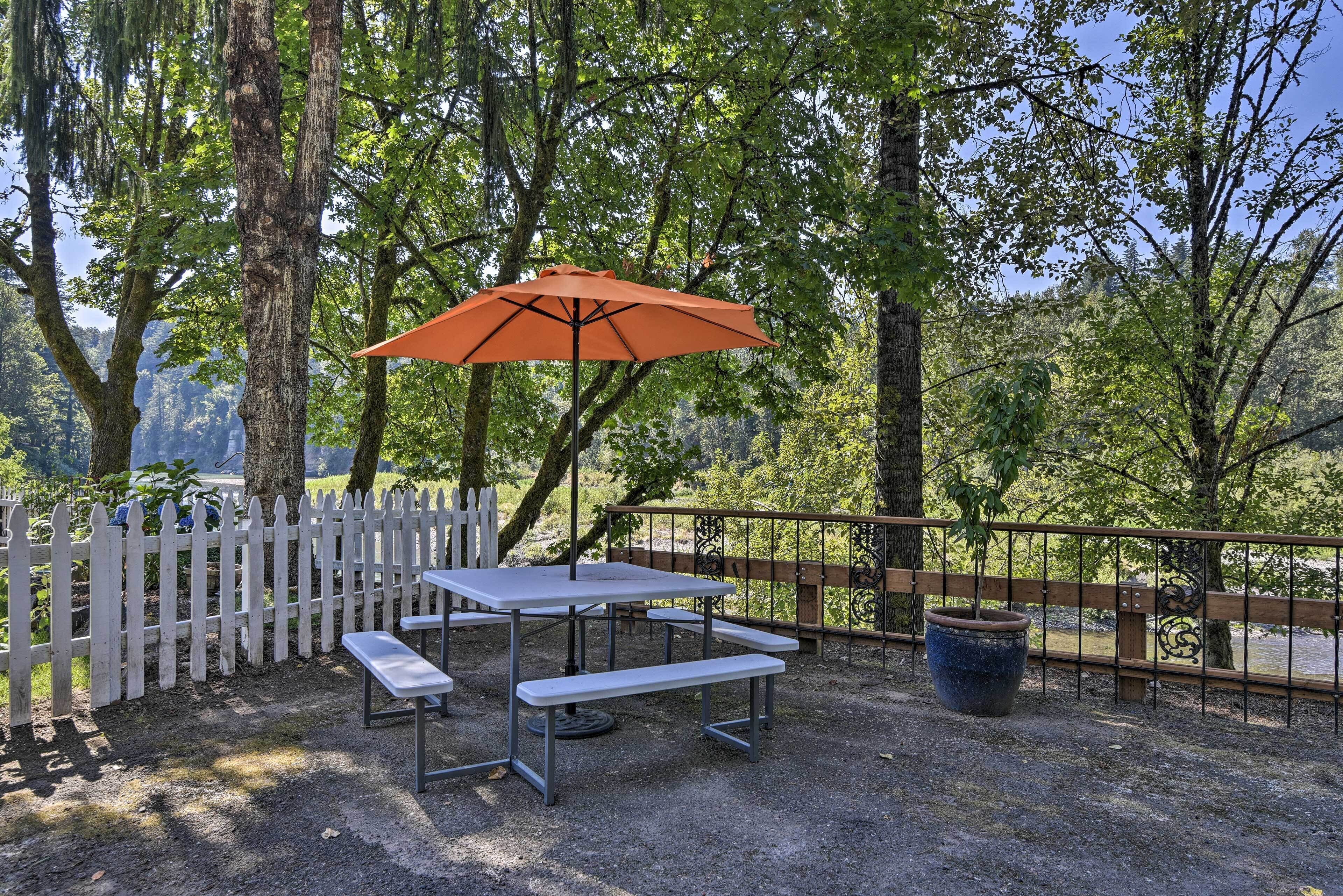 A picnic table with benches and an orange umbrella in a wooded area with a white picket fence.