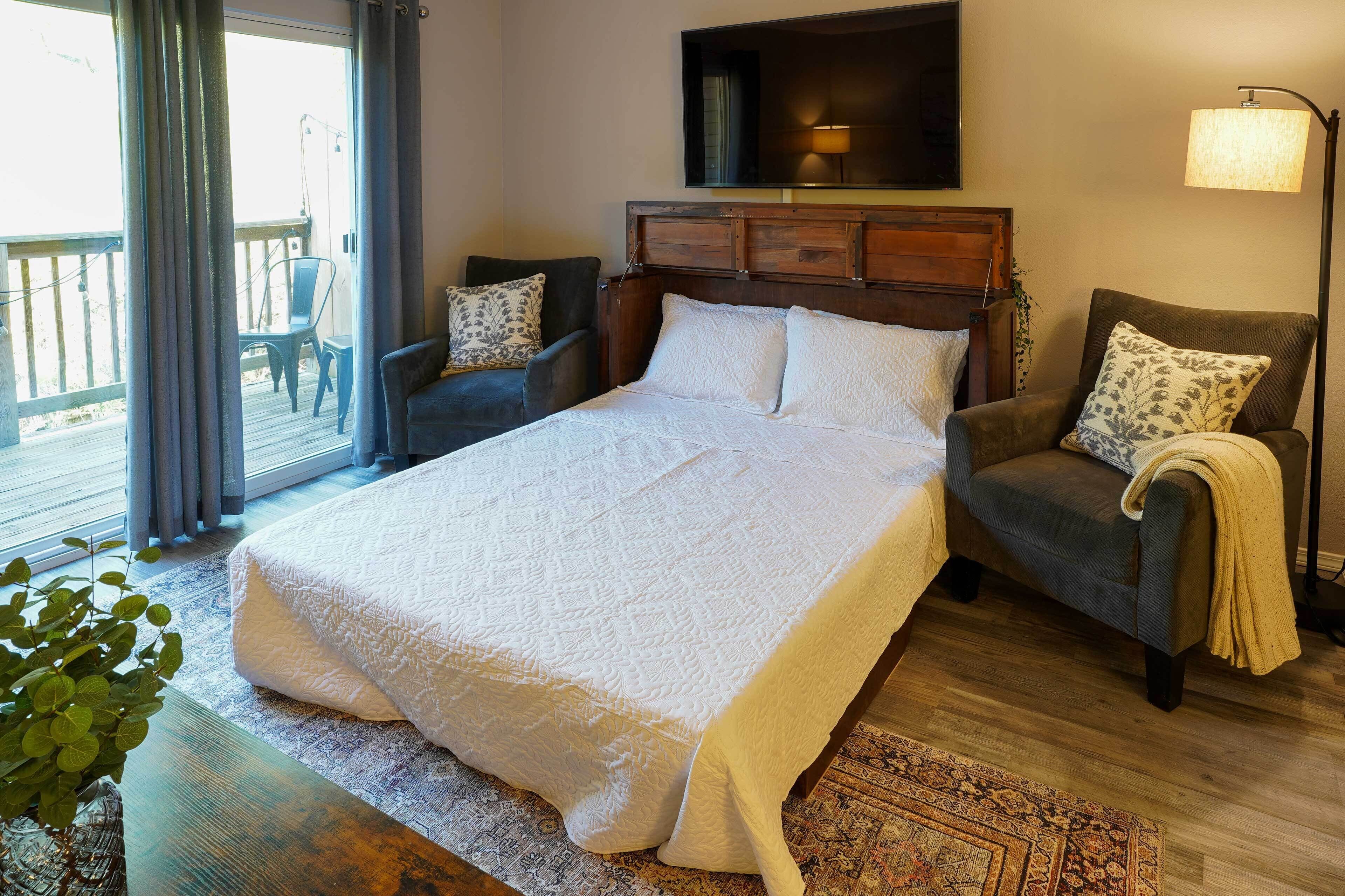 A bedroom with a large bed, two armchairs, a wooden headboard, a floor lamp, and a view of the outdoors.