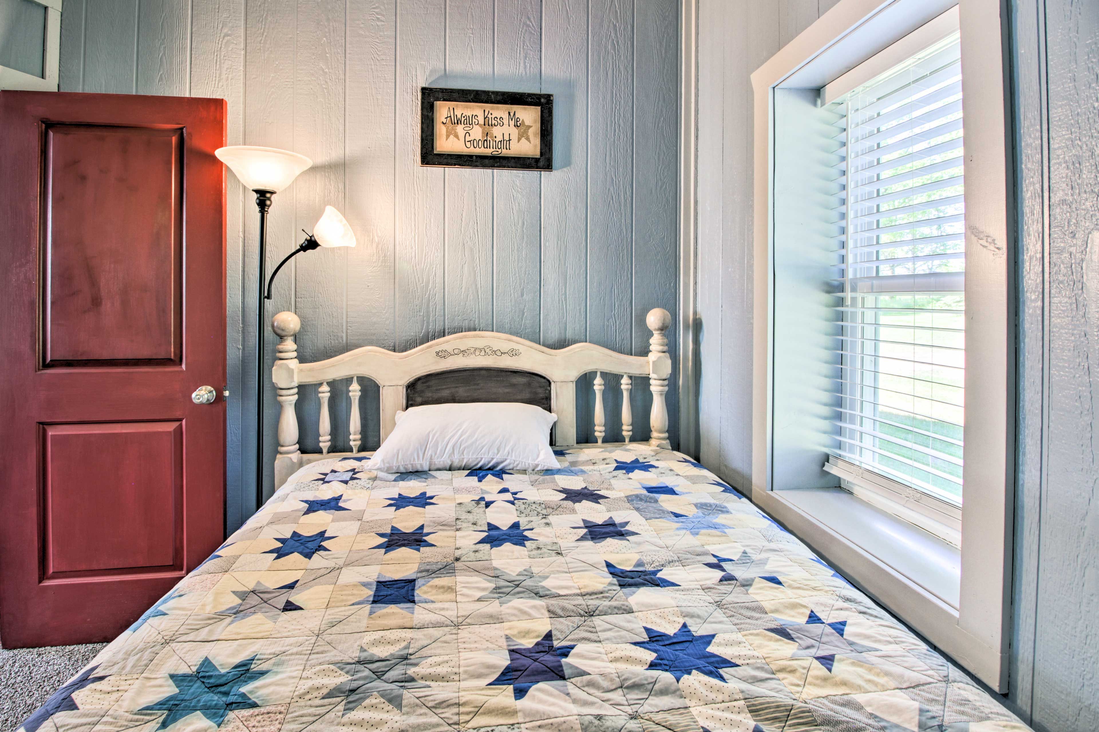 A bedroom with a wooden bed, a nightstand, a lamp, a window with blinds, and a wall-mounted sign.