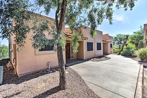 A residential building with a concrete driveway, a tree in the foreground, and a well-maintained garden with various plants and a cactus.
