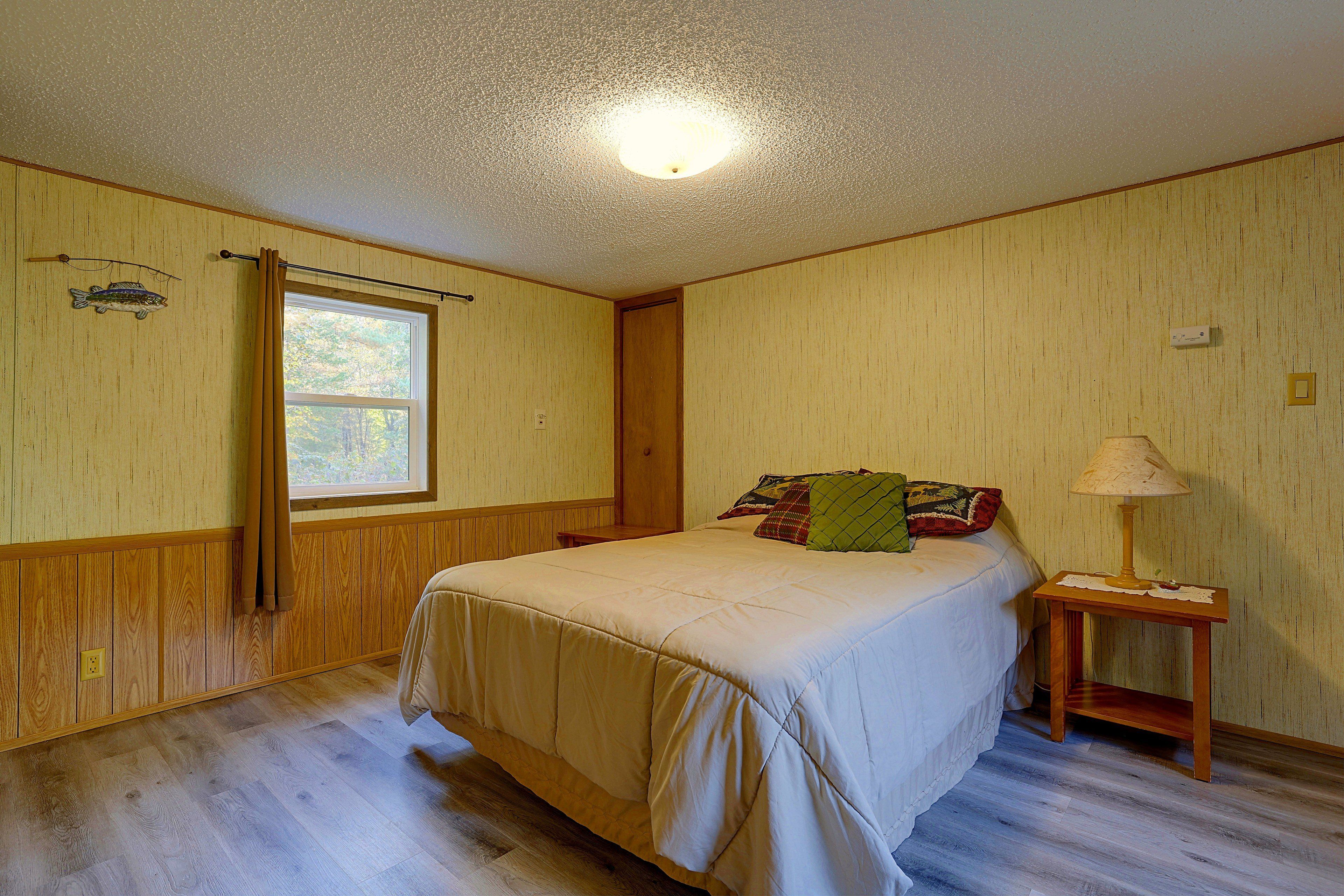 Quarto com cama, mesa de cabeceira, luminária, janela e termostato fixado na parede.
