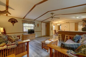 Uma sala de estar com televisão, móveis de madeira, ventilador de teto e área de cozinha com geladeira e micro-ondas.