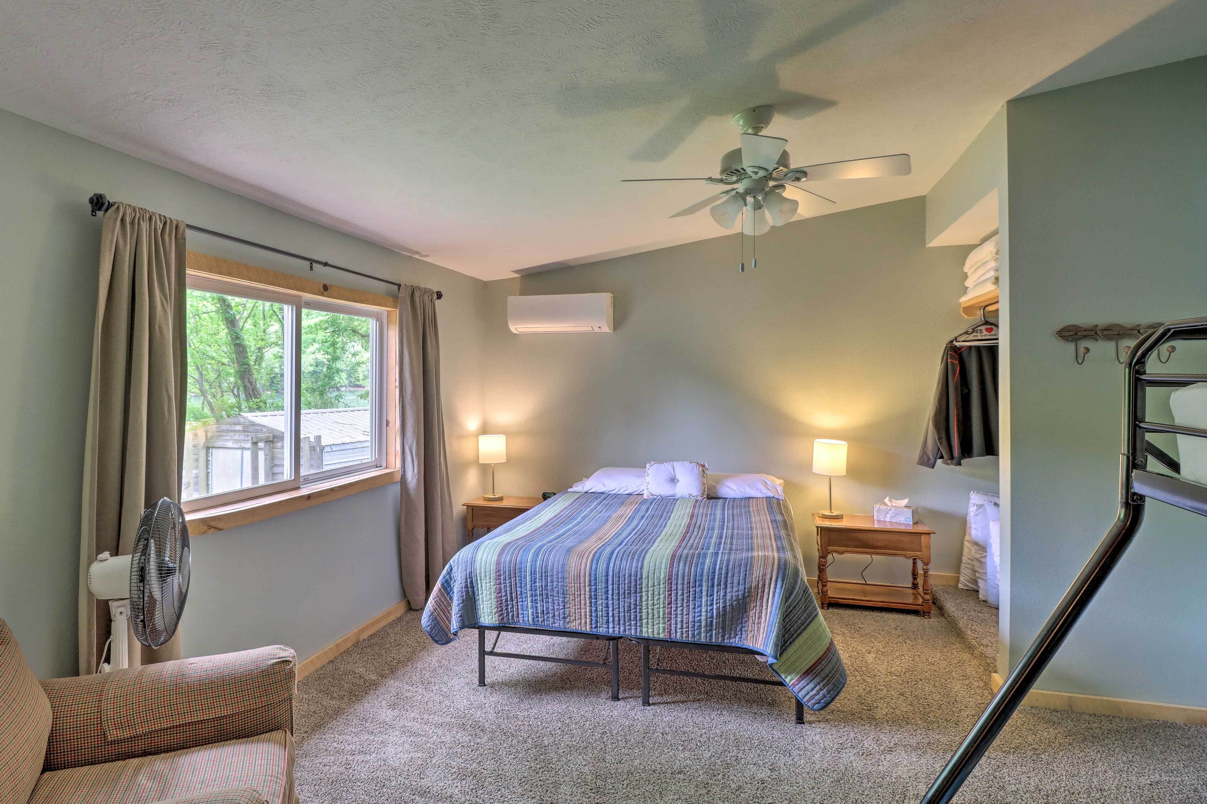 A bedroom with a bed, a ceiling fan, a window with curtains, a bedside table with a lamp, and a chair.