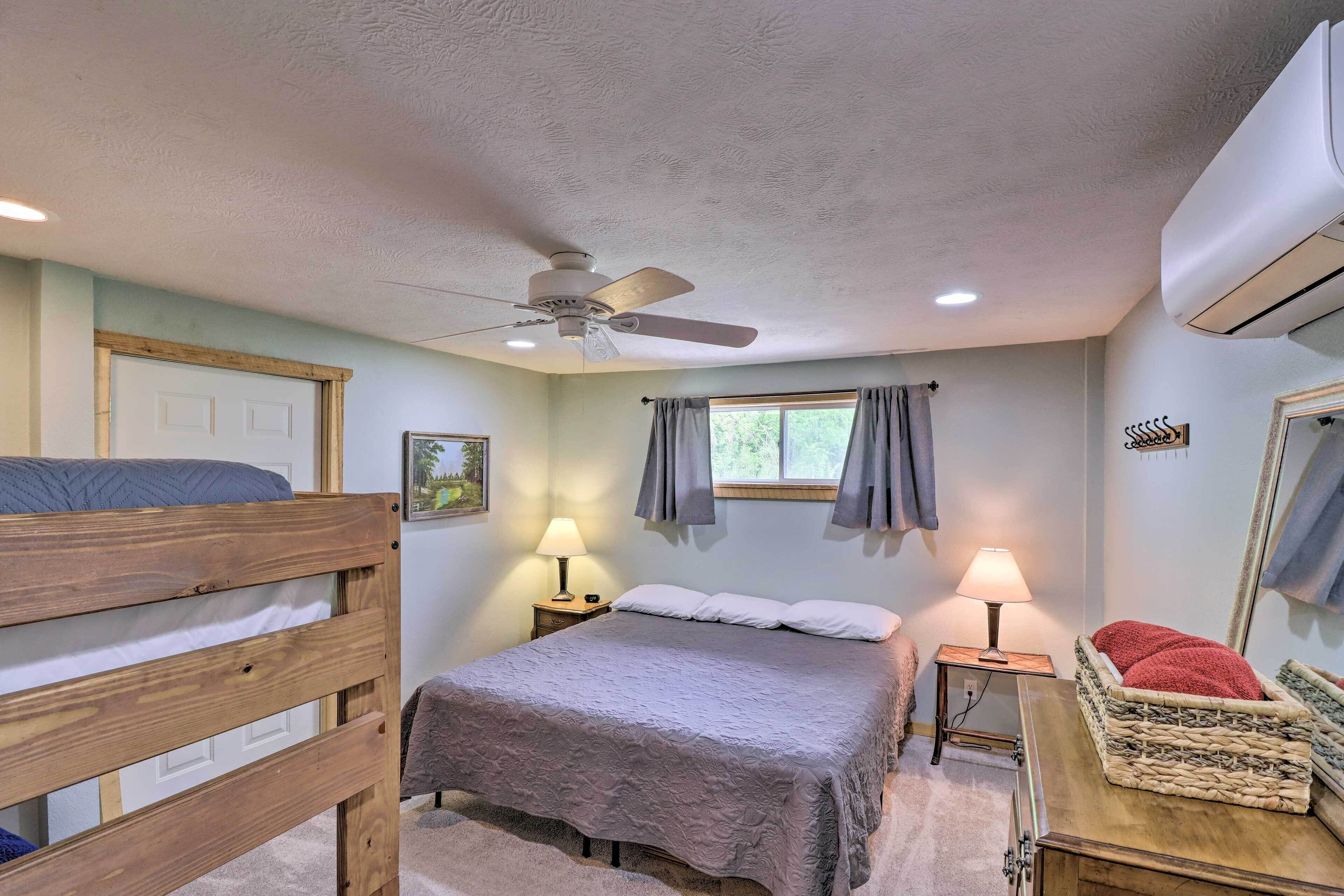 Um quarto com cama beliche, uma cama de solteiro, ventilador de teto, ar condicionado, duas luminárias de mesa, janela com cortinas e uma mesinha com um cobertor vermelho.