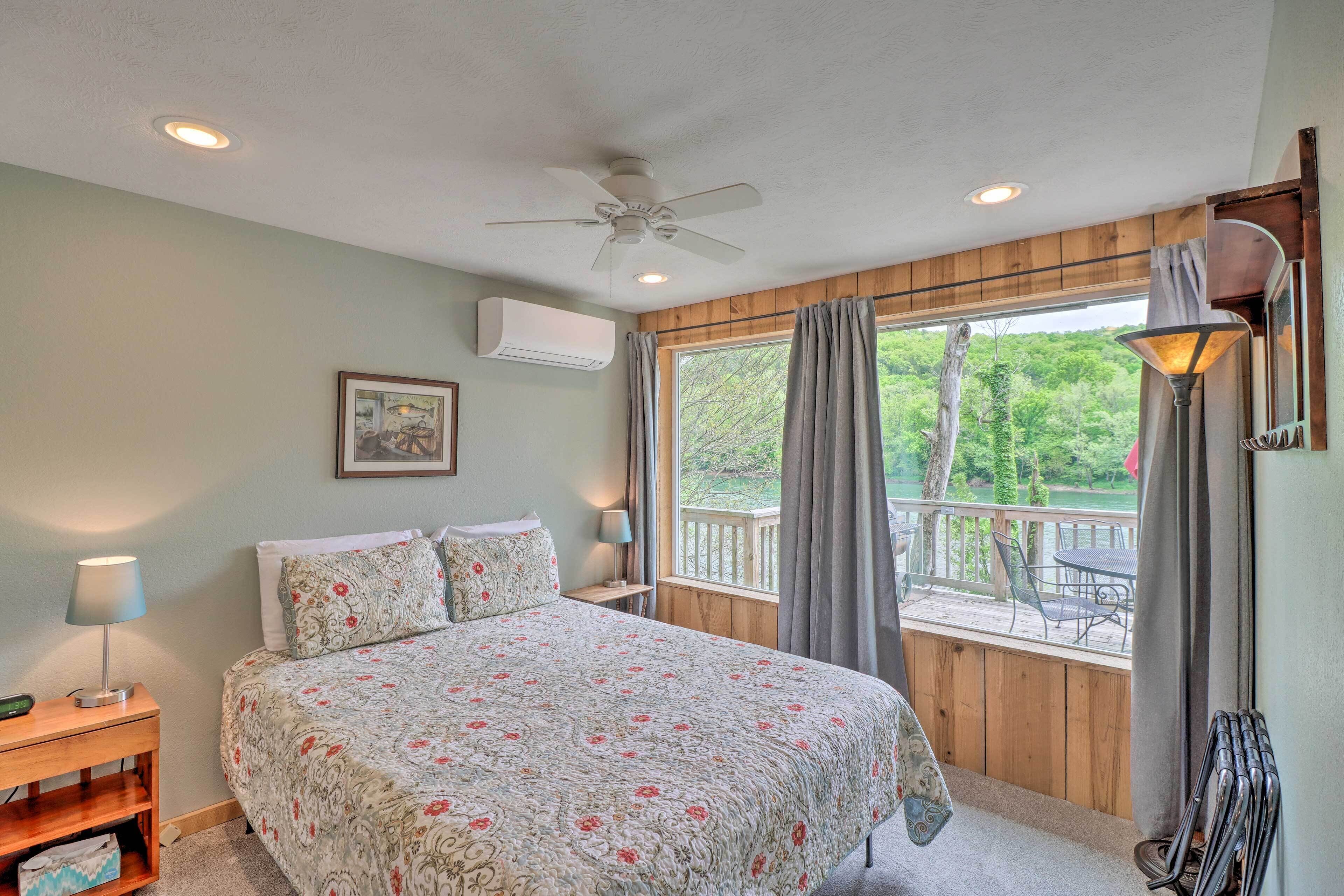 A bedroom with a bed, a ceiling fan, an air conditioner, a nightstand, a lamp, a window with curtains, and a view of the outdoors.