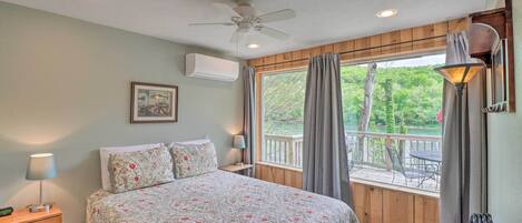 A bedroom with a bed, a ceiling fan, an air conditioner, a nightstand, a lamp, a window with curtains, and a view of the outdoors.