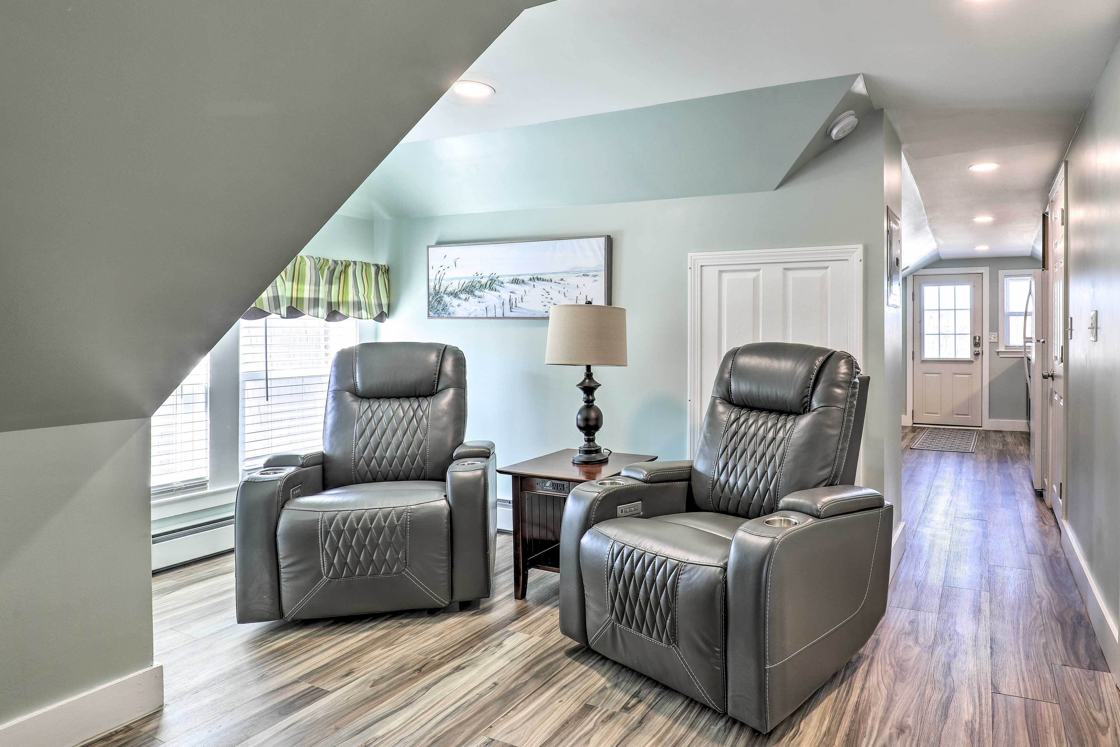 A modern living room with two leather recliners, a small table with a lamp, and a view of a hallway with a door and a window.