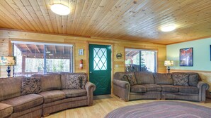 A cozy living room with wooden ceilings and walls, featuring two sectional sofas with patterned cushions, a green door, and a large window with blinds. - Near Natl Parks! Secluded Duck Creek Village Gem (Duck Creek Village)