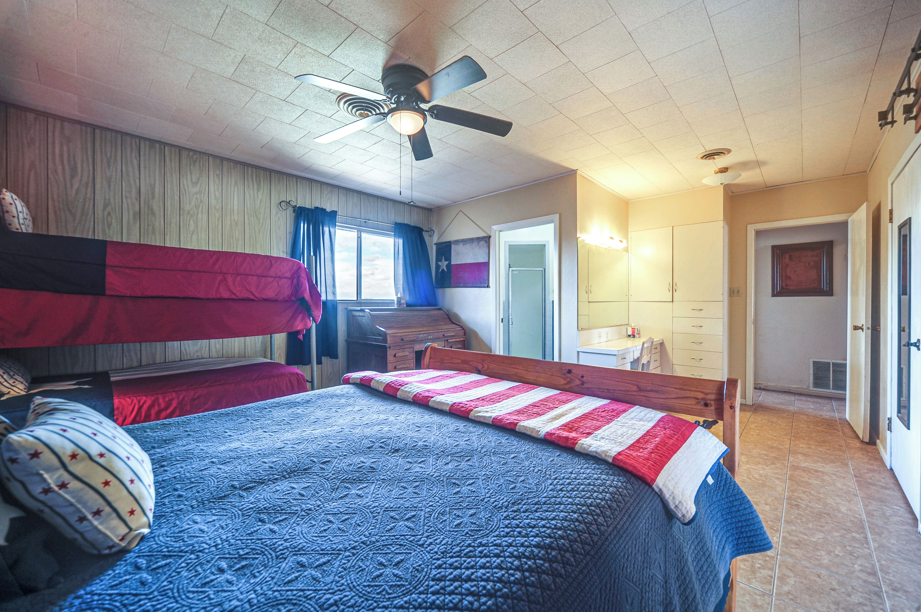 Ein Schlafzimmer mit Etagenbetten, Deckenventilator, Fenster mit Vorhängen, Schreibtisch und einem Badezimmer mit Dusche und Toilette.