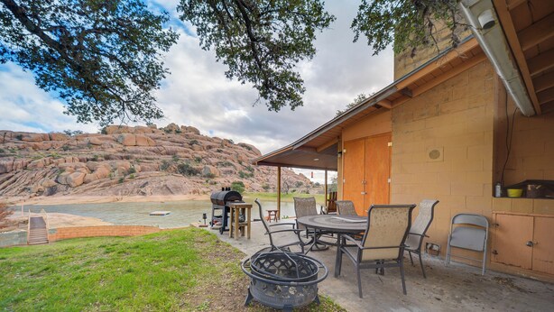 An outdoor patio with a fire pit, dining table, and chairs, overlooking a river and rocky hills.