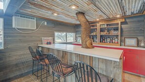 A rustic bar area with a wooden bar counter, metal chairs, and a unique tree trunk centerpiece.