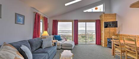 A spacious living room with a grey sofa, a wooden coffee table, and a dining area with a wooden table and chairs. There are large windows with red curtains, a view of the outdoors, and a ceiling with skylights.