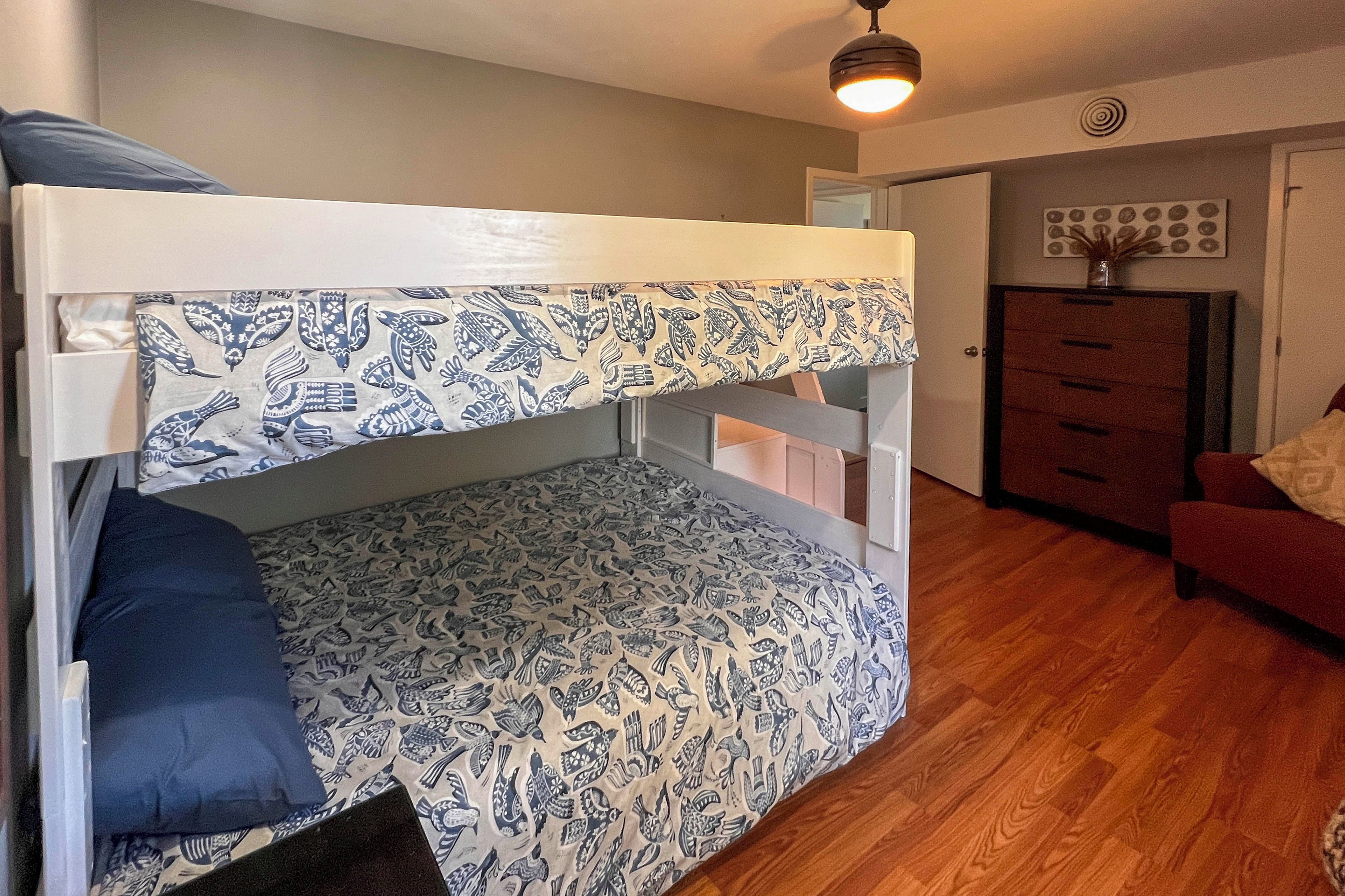 A bunk bed with patterned bedding, a wooden dresser, and a red sofa in a room with wooden flooring.