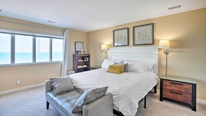 A bedroom with a large bed, a nightstand, a lamp, and a view of the ocean. - Lakefront Family Retreat w/ Grill: Steps to Beach! (Gary)