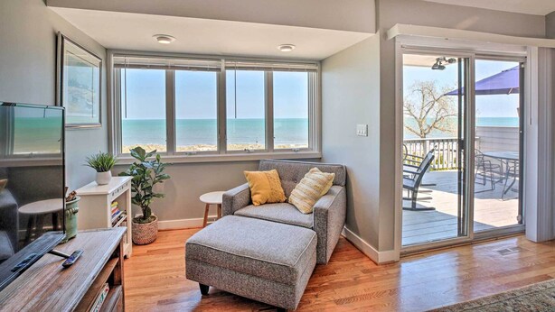 A living room with a gray sofa, a coffee table, a TV, and a view of the ocean through large windows. - Lakefront Family Retreat w/ Grill: Steps to Beach! (Gary)