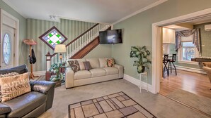 Un salon avec un escalier, un téléviseur fixé au mur et un coin repas visible à travers une porte ouverte.