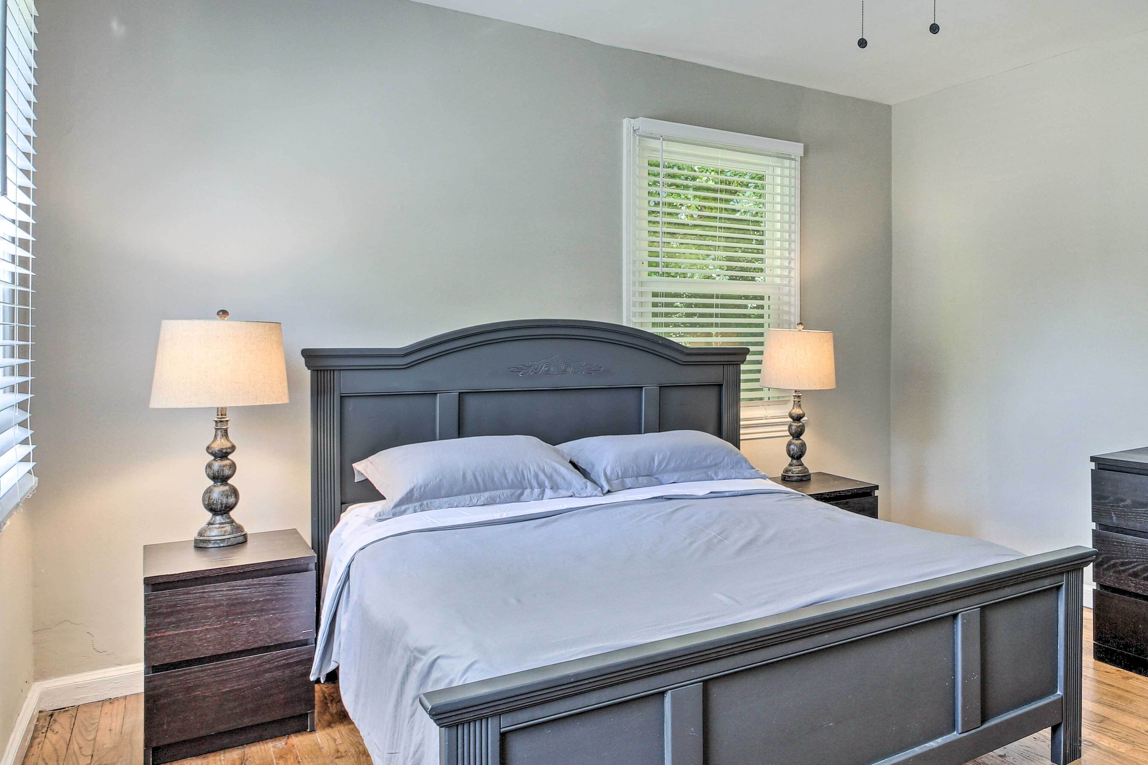 A bedroom with a bed, two bedside tables with lamps, a window with blinds, and a dark headboard.