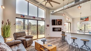 A modern living room with a leather sofa, a wooden coffee table, and a large window with a view of a mountainous landscape. - Chic Sedona Retreat: 2 Mi to Cathedral Rock Trail! (Sedona)