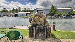 A stone fireplace with a metal door and a star design, set on a gravel area with a green chair and a dock extending into a lake. - Weirs Channel Cottage w/ Beach Access! (Laconia)