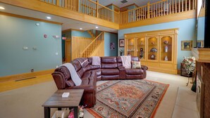 A spacious living room with a large brown leather sectional sofa, a wooden staircase, and a built-in cabinet with decorative items. - Hot Tub & Private Pond! Spacious Mcarthur Home (McArthur)