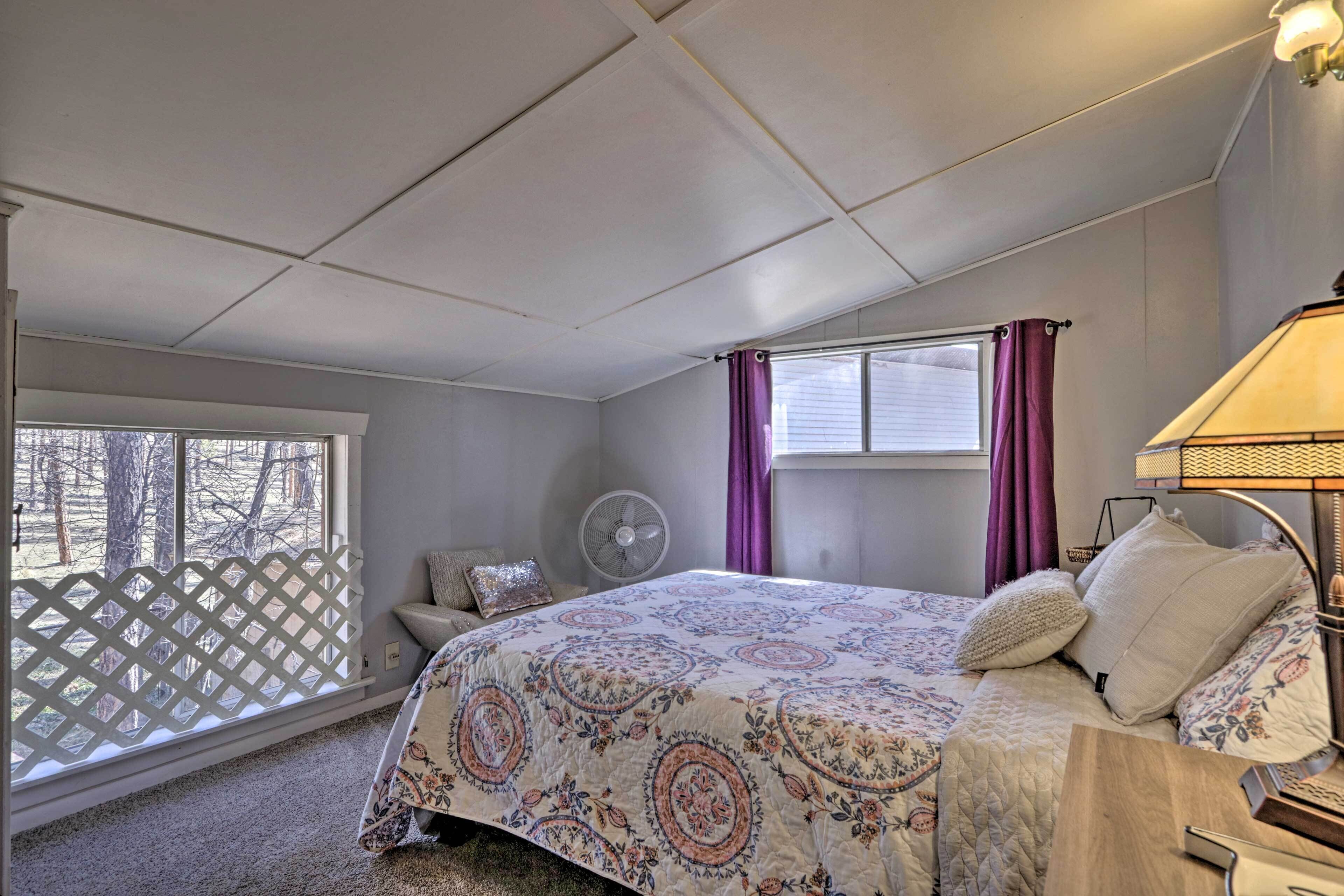 A bedroom with a bed, a nightstand, a lamp, a window with curtains, a fan, and a view of trees outside.