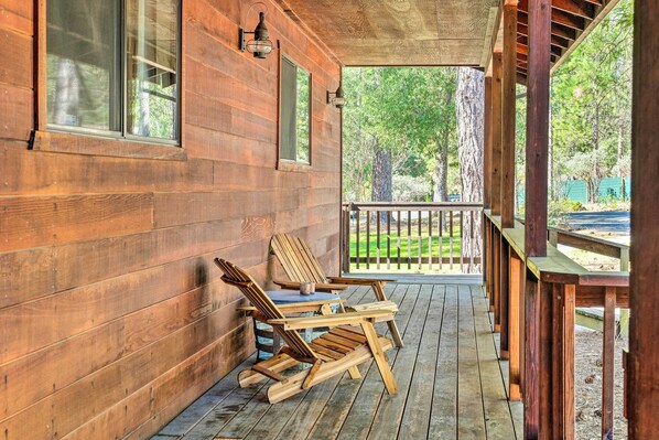 Eine hölzerne Veranda mit zwei Adirondack-Stühlen und einem Tisch, mit Blick auf eine Rasenfläche mit Bäumen.