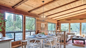 Une cabane en bois spacieuse, dotée de grandes fenêtres, d’un coin repas avec des chaises blanches et d’un espace de vie confortable avec un canapé et une table basse.
