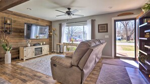 A modern living room with a large flat-screen TV, a beige recliner, wooden paneling, a ceiling fan, and a view of the outside through a glass door. - Quaint Creekside Cottage w/ Porch & Backyard! (Lexington)