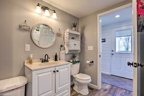 A bathroom with a white vanity, a toilet, and a sink. There is a mirror above the vanity, a light fixture above the mirror, and a door leading to a hallway.