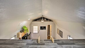 Un dormitorio en el ático con techo inclinado, un ventilador de techo, una puerta con ventana, un escritorio pequeño con una planta y una escalera que baja.