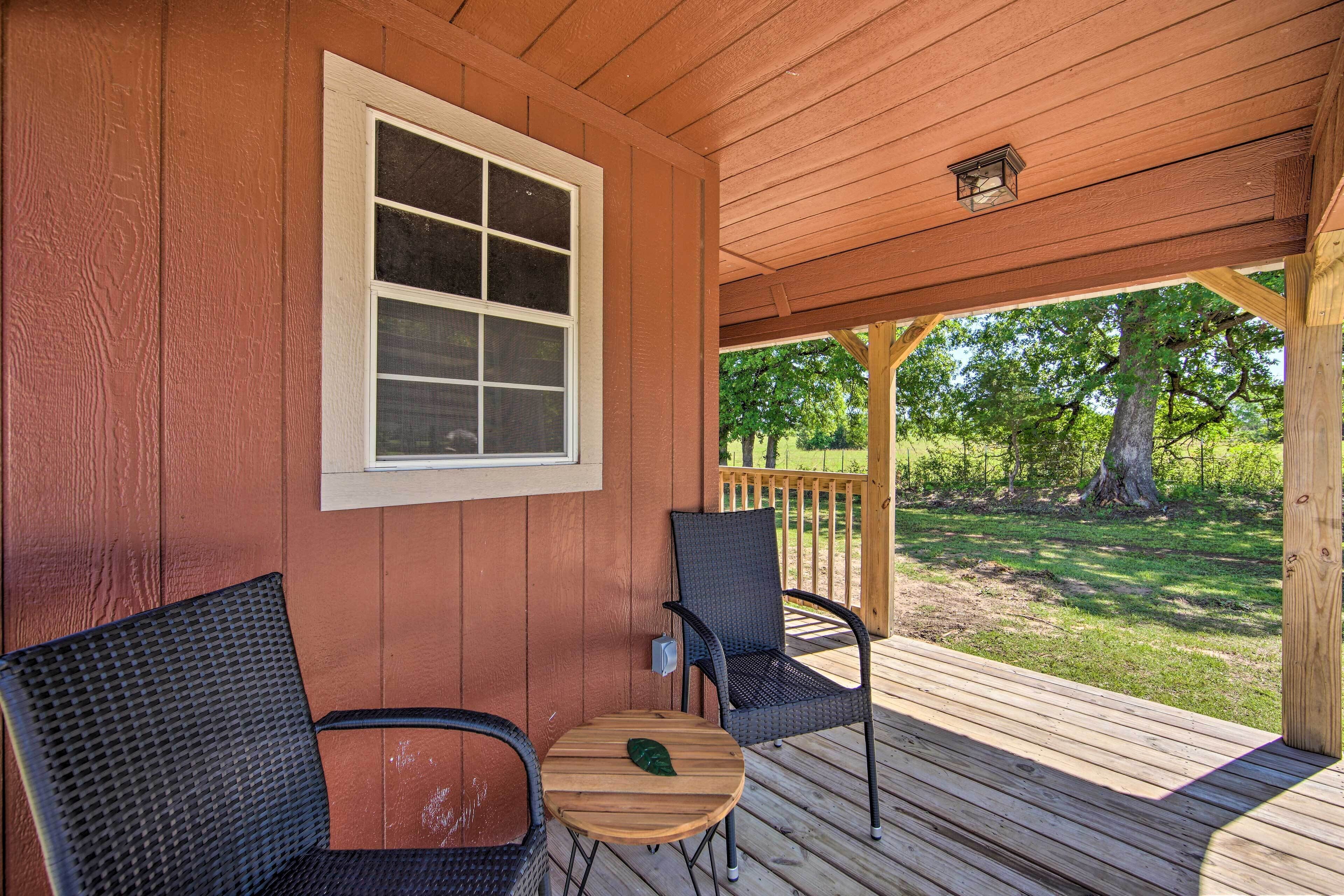 A wooden deck with two wicker chairs and a small table, a window, and a view of a grassy area with trees.