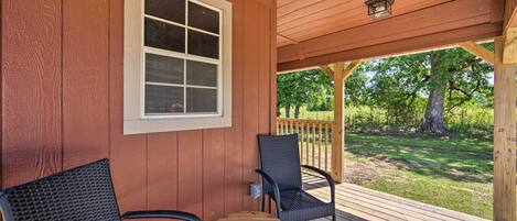 A wooden deck with two wicker chairs and a small table, a window, and a view of a grassy area with trees.