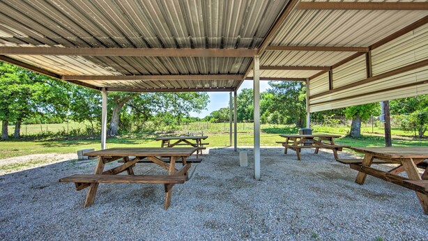 Un área de picnic techada con mesas y bancos de madera, suelo de grava y árboles al fondo.