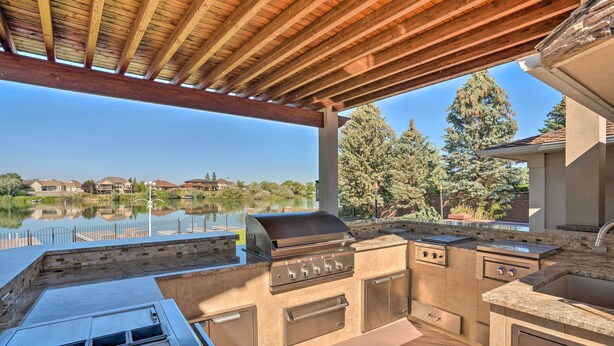 Cocina exterior con parrilla, pileta y alacenas, con vista a un lago y casas al fondo.