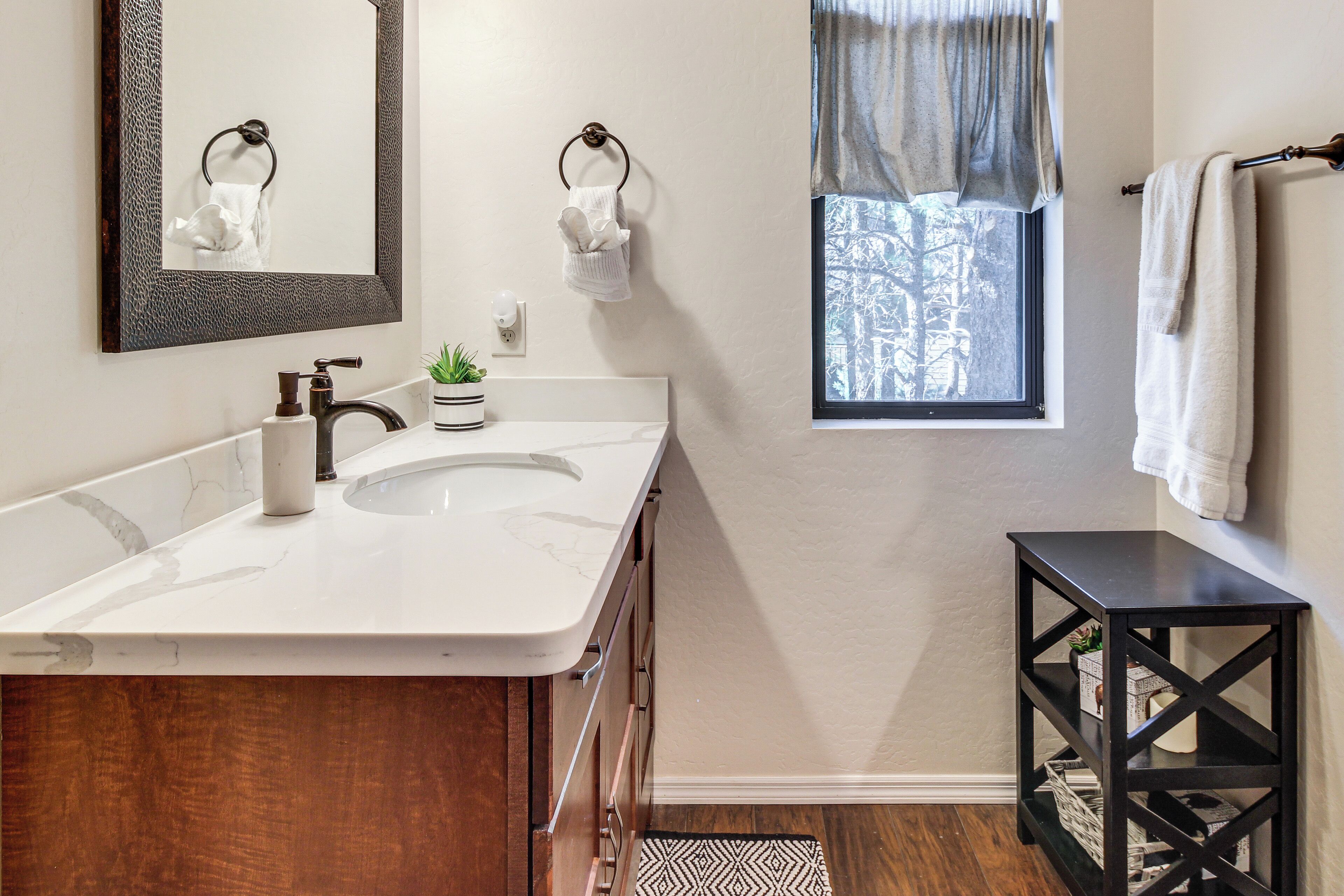 A bathroom with a white sink, dark wooden vanity, a mirror with a textured frame, a window with a view of trees, and a black shelf with a plant.